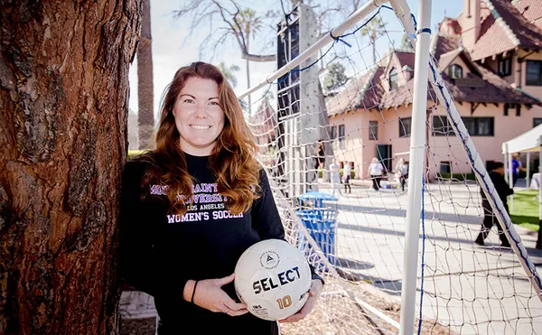 A student holding a soccer ball and leaning against a tree with a soccer goal and MSMU Doheny buildings in the background.