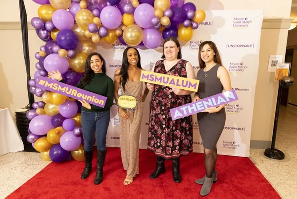 four women smiling in front of purple and gold MSMU backdrop holding signs