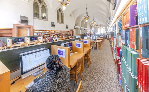 A row of compter desks in a large library hall with a student at one of the desks