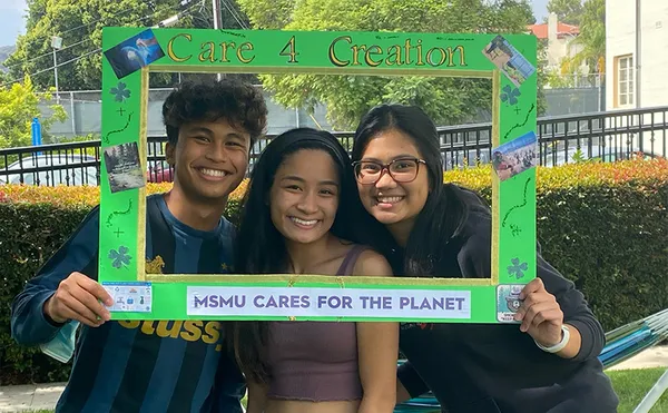 Three students holding up and smiling from behind a decorated green picture frame with the words: 