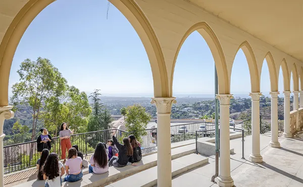 A view of students sitting on steps and listening to another student standing in front of them seen through an arched walkway