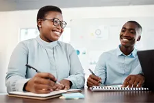 two people smiling working together at a table with notepads