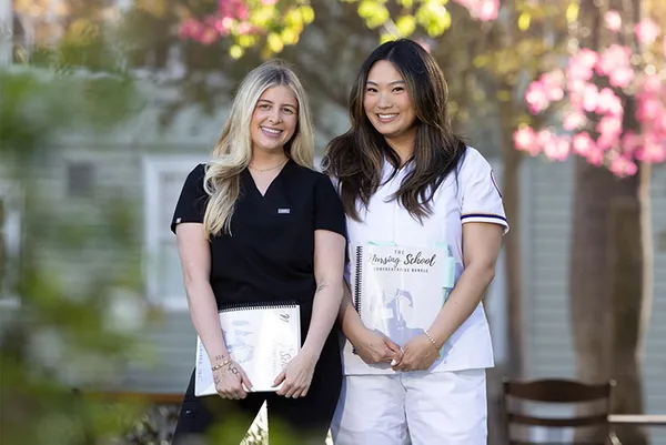 two female students smiling on campus, holding nursing booklets