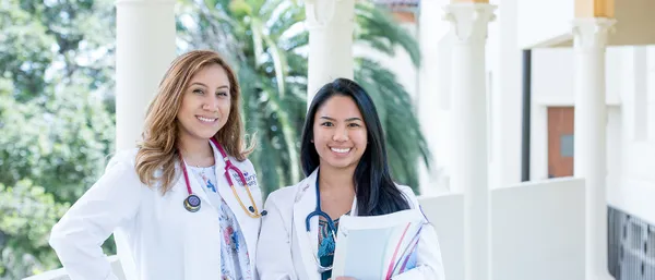 two female nursing students smiling while standing outdoors and wearing white lab coats
